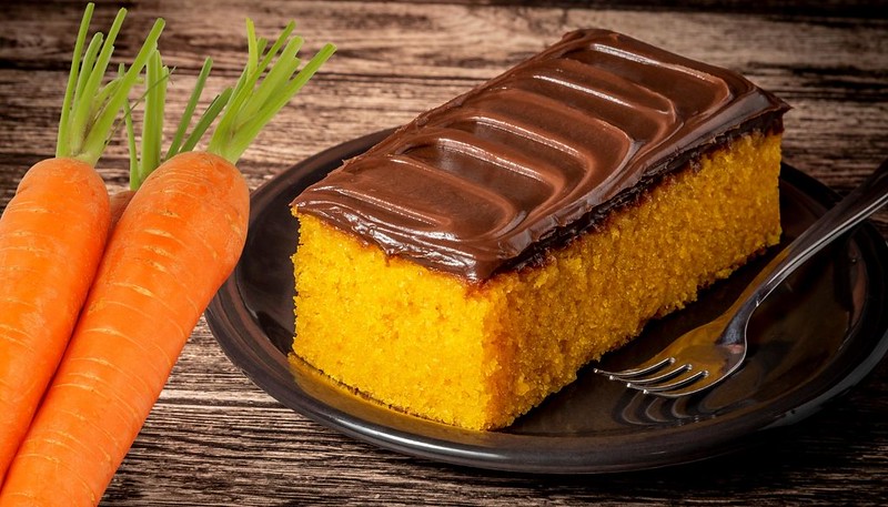 Pedaço de bolo feito com cenoura com cobertura de chocolate, em um prato com um garfo do lado, e duas cenouras do outro lado.