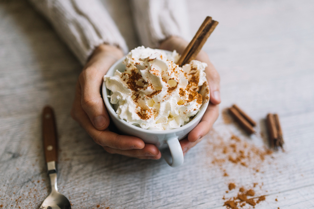 Duas mão segurando uma xícara com bebida quente e chantilly por cima, com umpouca de anela em pó.